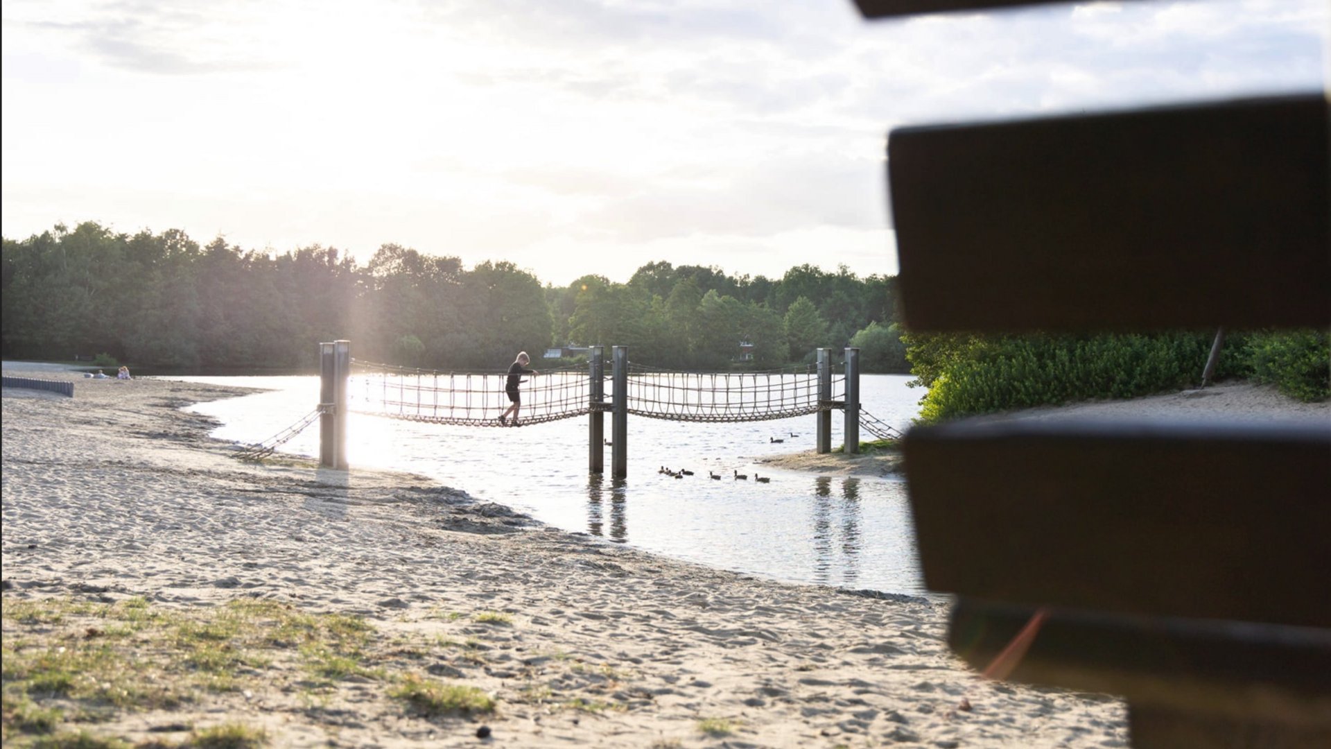 Erholung, Abenteuer und Badespaß für die ganze Familie – direkt am Dreiländersee Kind auf einer Hängebrücke über einem See bei Sonnenuntergang
