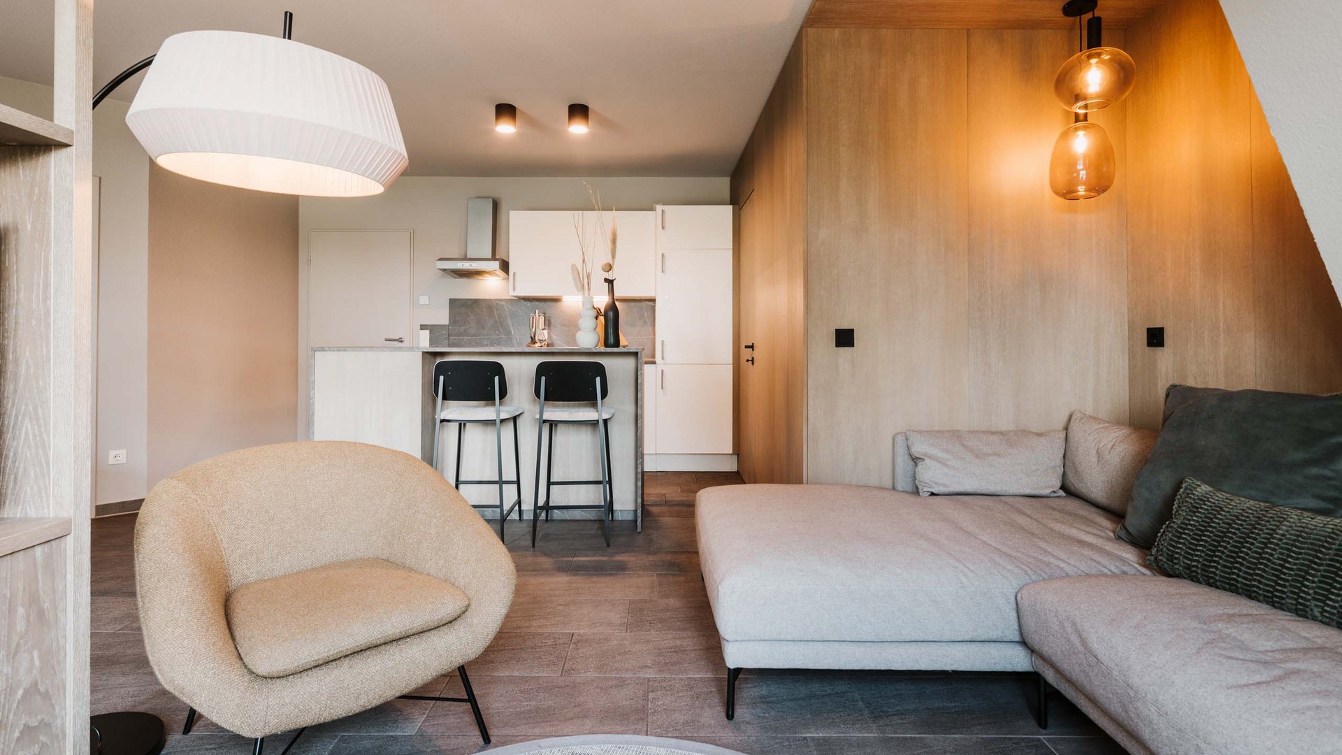 Unsere Appartements am See Modernes Wohnzimmer mit beigem Sessel, grauer Couch und Küche im Hintergrund