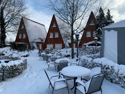 Urlaub am See in NRW - sogar im Winter wunderschön! Schneebedeckte Tische und Stühle vor drei spitzen Holzhäusern im Winter