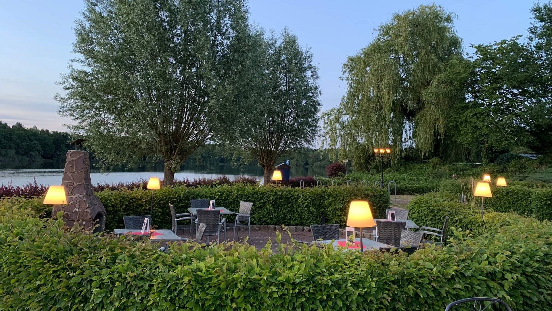 Jobs im Hotel Restaurant Seeblick in Gronau Abendliche Terrasse mit beleuchteten Tischen neben einem See und Bäumen