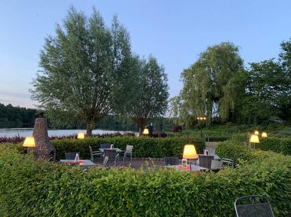Urlaub im Apartment mit Seeblick Abendliche Terrasse mit beleuchteten Tischen neben einem See und Bäumen