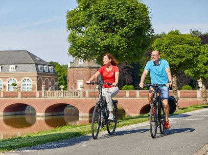 Seeblick Gronau: Urlaub inmitten der Natur Paar fährt bei Sonnenschein auf Fahrrädern an einem Schloss und Park vorbei