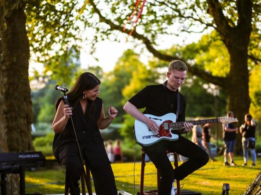 Seeblick Gronau: Urlaub inmitten der Natur Zwei Musiker spielen draußen Gitarre und singen unter Bäumen auf einer kleinen Bühne.